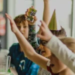 Children sit at a table enjoying a festive football birthday party with pizza and drinks. One child wears a party hat, and several have their hands raised excitedly. The atmosphere is lively and joyful. © Powerleague