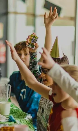Children sit at a table enjoying a festive football birthday party with pizza and drinks. One child wears a party hat, and several have their hands raised excitedly. The atmosphere is lively and joyful. © Powerleague