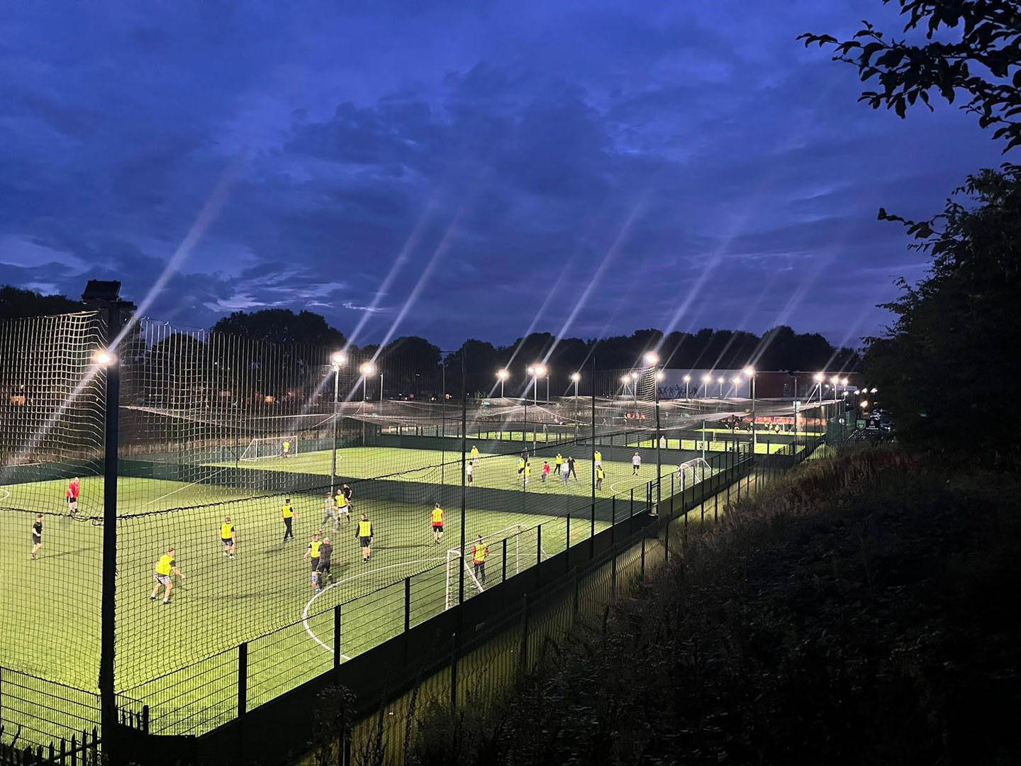 A nighttime scene of multiple illuminated soccer fields surrounded by tall fencing. Players in bright jerseys are actively participating in games, representing local football leagues on the fields. The sky is a deep blue with scattered clouds, and trees line the background beyond the fields. © Powerleague