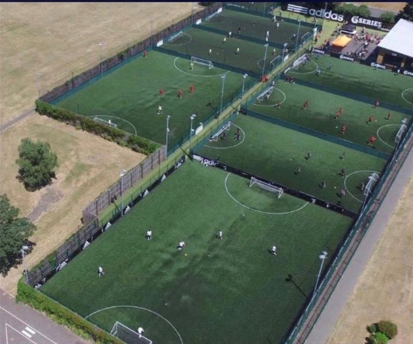 Aerial view of multiple small, fenced soccer fields bustling with local football leagues in action. The surrounding area features lush grass and trees. A building boasting an Adidas logo is visible in the top right corner. © Powerleague