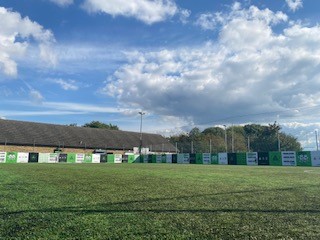 A grassy field hosts a row of green and white banners along a fence, hinting at local football leagues. A building peeks out behind the banners, while trees reach up to meet the blue sky dotted with clouds. © Powerleague