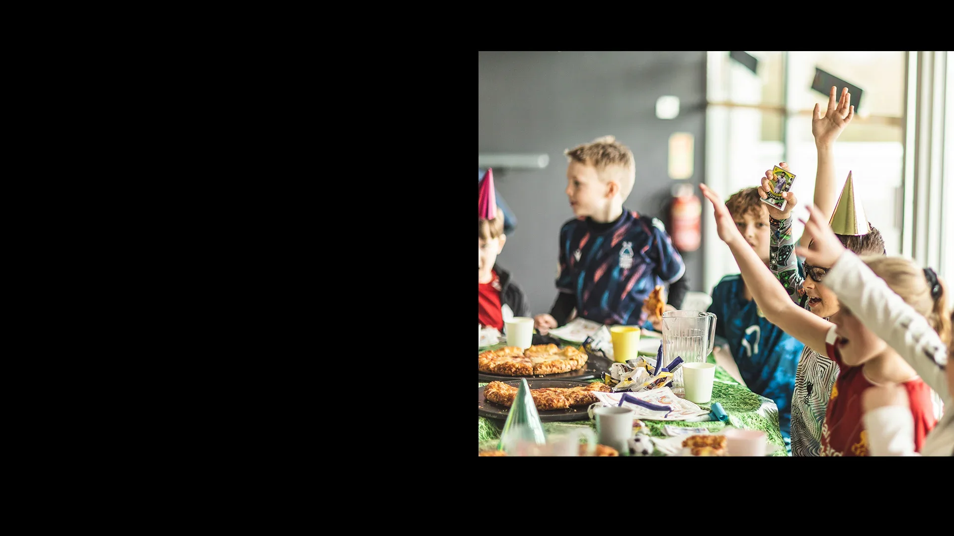 Children wearing party hats are sitting at a table decorated with a green tablecloth, filled with various food items including pizzas and desserts. The excitement is palpable at the football birthday party, with one child enthusiastically raising their hand. The background shows a blurred indoor setting. © Powerleague