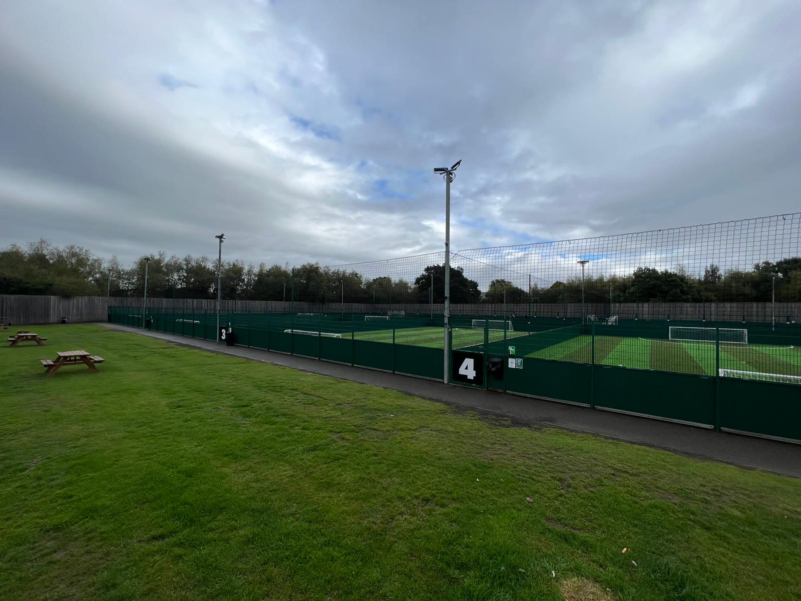 An outdoor sports complex with several fenced artificial turf soccer fields hosts local football leagues under a cloudy sky. Numbered signs are visible on the fences, and the foreground shows grassy areas with picnic tables to the left. © Powerleague