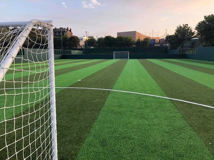 A soccer field with green and darker green striped artificial turf hosts local football leagues. A goalpost and net are in the foreground, with another in the distance. The sky is clear, and trees and buildings dot the background, setting a perfect scene for grassroots competition. © Powerleague