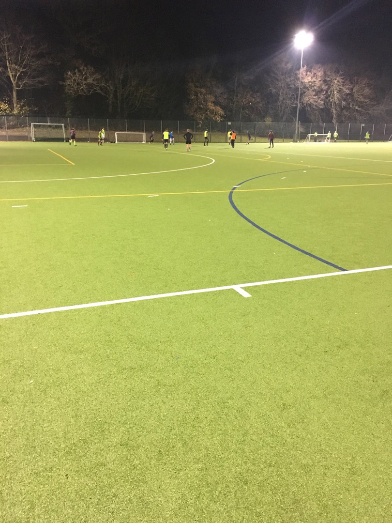 A night view of a soccer match in progress on a well-lit artificial turf field. Players in neon and dark uniforms, representing local football leagues, are scattered across the field, with a goalpost visible in the background. Trees surround the field, and shadows are cast by the bright stadium lights. © Powerleague