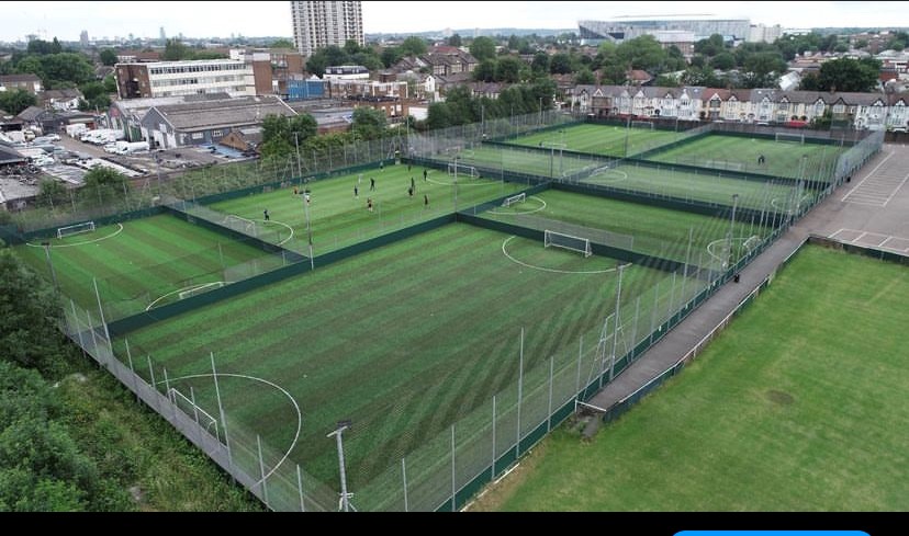 Aerial view reveals multiple fenced soccer fields with vibrant green artificial turf, hosting players likely from local football leagues. Surrounding the pitches are residential houses, industrial buildings, and a large sports stadium looming in the background. © Powerleague