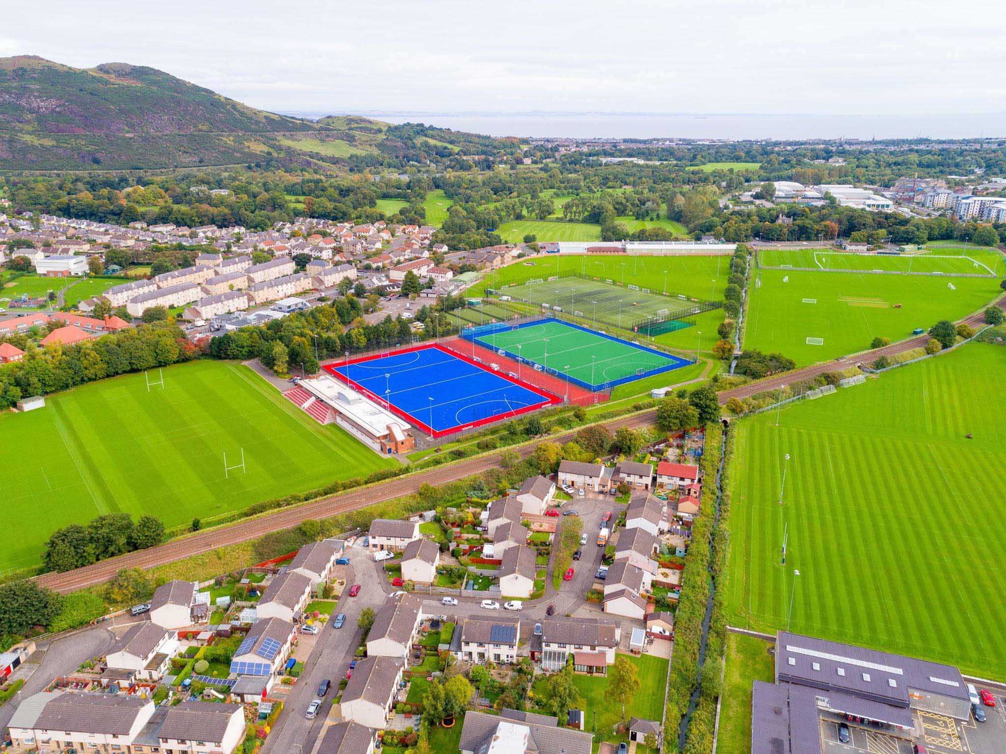Aerial view of a sports complex, where local football leagues play on vibrant blue and green fields for hockey, enclosed by red and blue fences. The surrounding residential area features houses, roads, lush greenery, and hills in the background under a clear, bright sky. © Powerleague