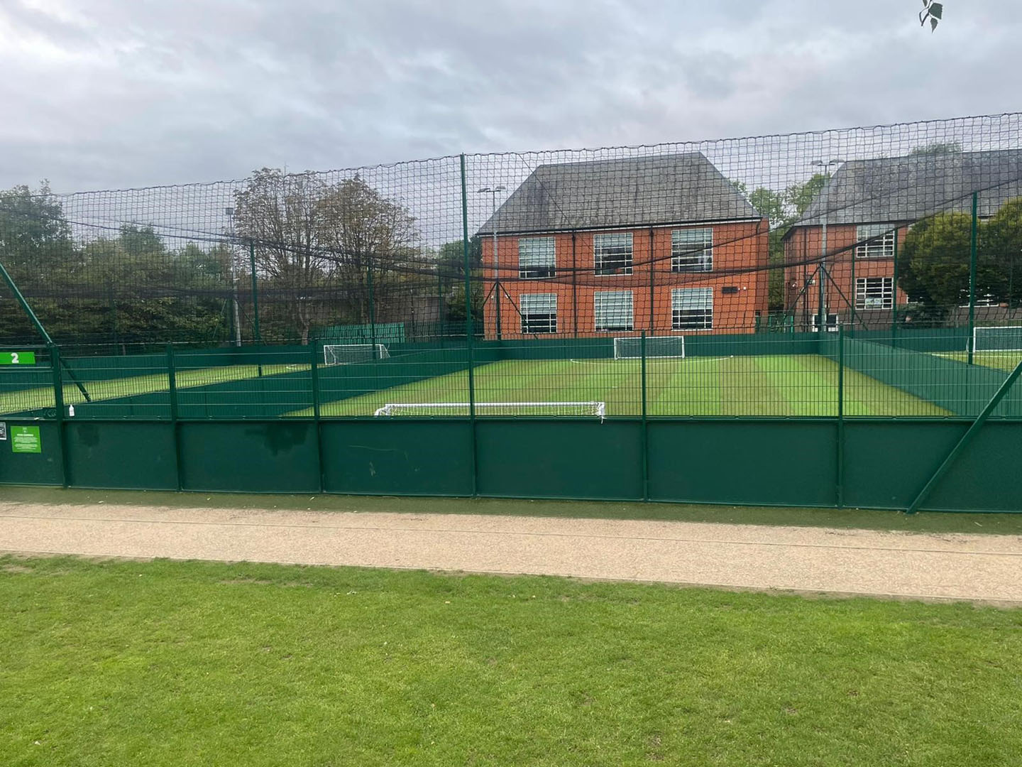 A fenced outdoor sports field with artificial grass, featuring two small soccer goals, serves as a lively venue for local football leagues. The background showcases a red brick building with several windows and a gray roof. The sky is overcast, with a grassy area in the foreground adding charm. © Powerleague