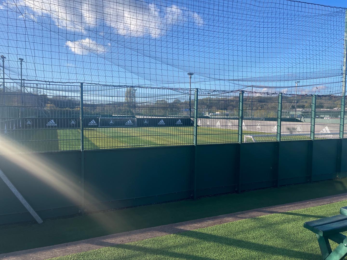 A fenced outdoor sports field, often hosting local football leagues, stretches under a clear blue sky. Adidas logos line the inner fence with pride. Sunlight streams from the left, casting a bright glare on the green artificial turf covering the foreground. © Powerleague