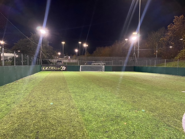 A well-lit, outdoor soccer field at night, perfect for local football leagues. The artificial turf is green, with a goal visible in the background. Tall lights line the sides, and trees can be seen outside the fenced area. © Powerleague