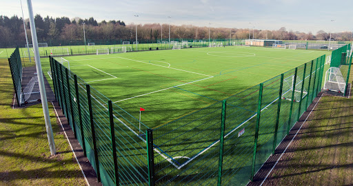 A large, artificial turf sports field hosts local football leagues, surrounded by a tall green fence. The field is marked with white and red lines for various sports and has goals on either end. Trees and a clear blue sky paint the backdrop. © Powerleague