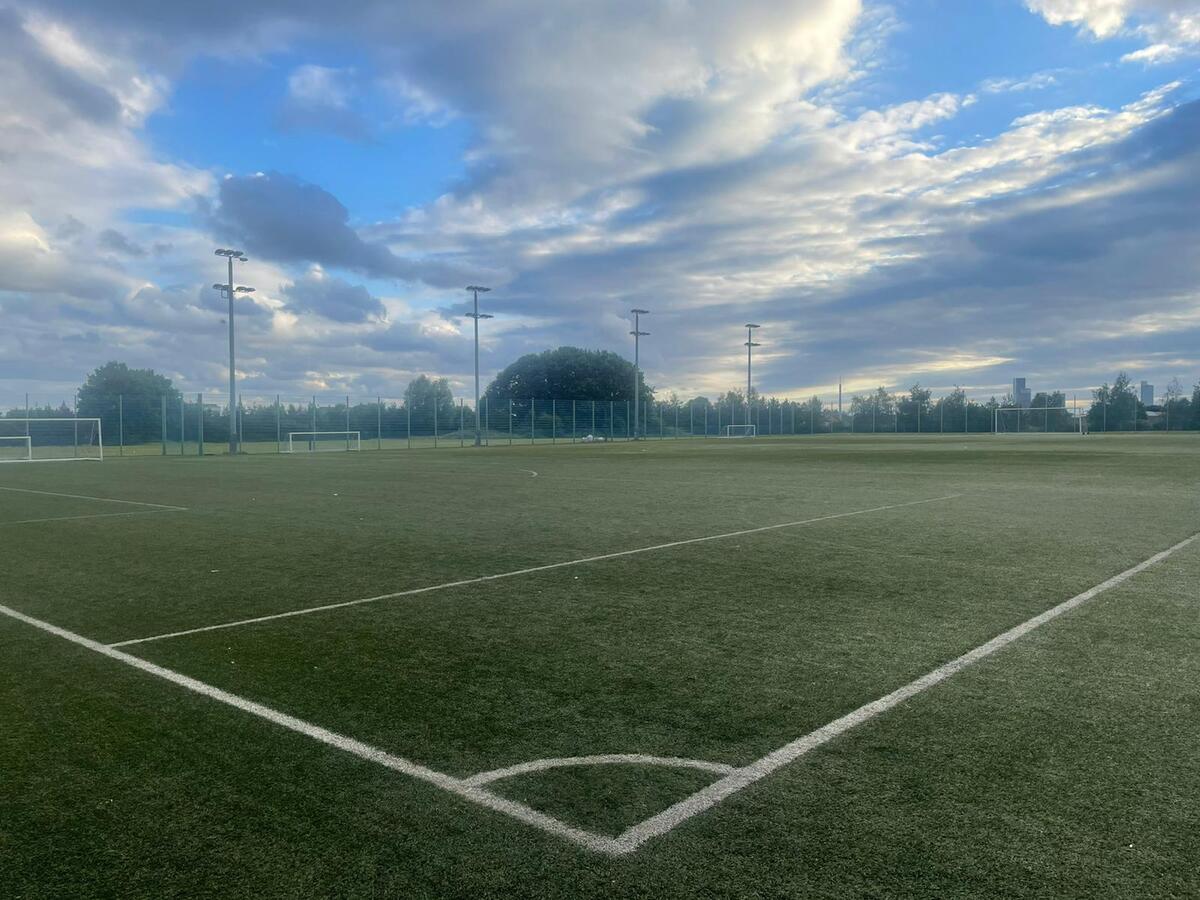 A photograph of an empty soccer field under a partly cloudy blue sky. The artificial turf field, used by local football leagues, has white boundary lines and several goalposts in the distance. Trees and tall streetlights surround the field, with a city skyline visible on the horizon. © Powerleague