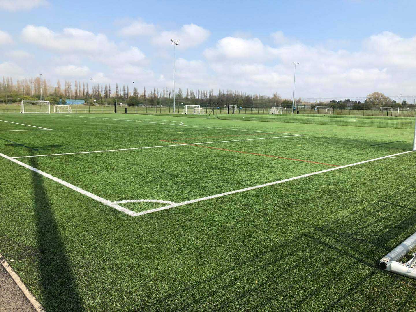 A well-maintained outdoor football field with bright green artificial turf and white boundary lines. Multiple goalposts can be seen in the distance under a partly cloudy blue sky, perfect for local football leagues. Trees and a fence line the background. © Powerleague