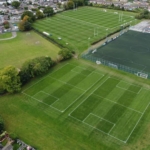 An aerial view of a sports complex showcasing multiple soccer fields with marked boundaries, often bustling with local football leagues. The fields are surrounded by trees, residential buildings, and parking lots, with some smaller structures visible within the area. © Powerleague