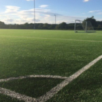 A well-maintained, green artificial turf soccer field is shown under a blue sky with scattered clouds. White boundary lines and a soccer goal are visible in the background, with trees and shrubbery lining the perimeter of the field, perfect for local football leagues. © Powerleague