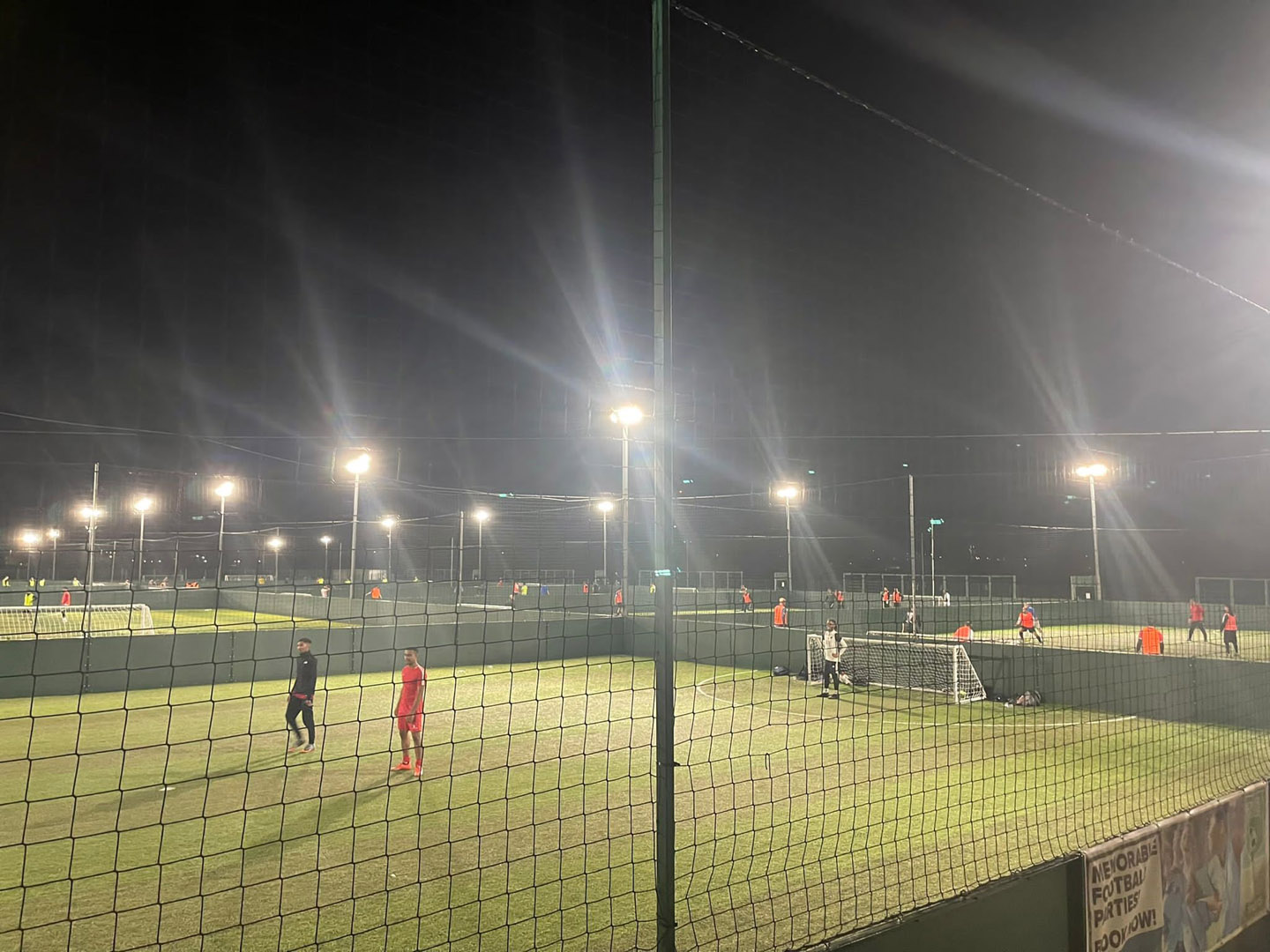 A nighttime soccer match from local football leagues is taking place on multiple illuminated fields, with players in jerseys actively playing. The fields are enclosed by netting, and bright floodlights cast a glow over the scene. © Powerleague