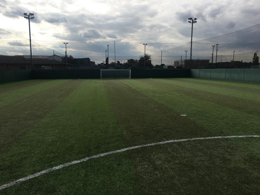 A small, enclosed soccer field with artificial turf under a cloudy sky serves as the perfect venue for local football leagues. The field is marked for gameplay, and a single goal stands at one end, illuminated by floodlights positioned around the perimeter. © Powerleague