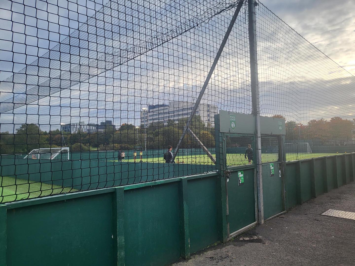 A fenced football field with artificial turf is shown, where local football leagues often play. Two people are walking near the gate, while trees and a building stand in the background under a cloudy sky. © Powerleague