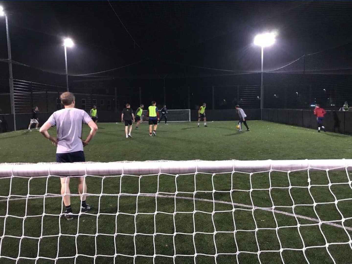 A group of people are playing soccer at night on a well-lit outdoor turf field. Viewed from behind a goal net, some players wear green pinnies, while others wear regular sports attire. The energetic scene is part of the local football leagues, with the game in progress under overhead lights. © Powerleague