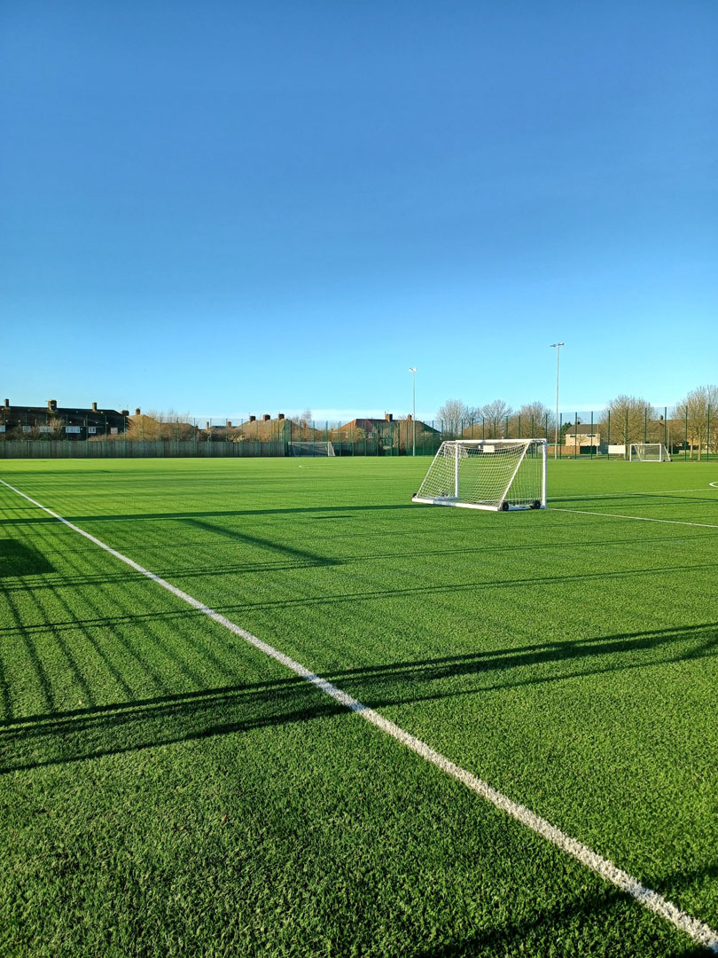 A spacious soccer field with artificial green turf, two small goals, and a clear blue sky hosts local football leagues. The field is surrounded by a fence, and residential buildings are visible in the background. © Powerleague